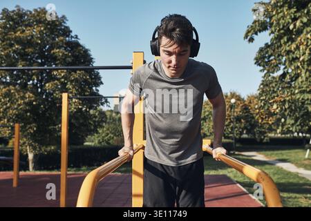 Giovane uomo che fa tuffi sulle barre parallele durante il suo workout in un parco calisthenics di strada moderno di workout. Uomo che ascolta musica indossando cuffie e Foto Stock