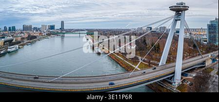Ampia vista aerea del ponte UFO a Bratislava che attraversa il Danubio con il lungofiume e lo skyline urbano; ideale per viaggi, trasporti, editoriale, touri Foto Stock