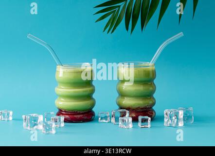 Due bevande di matcha con cannucce di vetro, cubetti di ghiaccio e foglie di palma tropicali su sfondo azzurro. Concetto creativo di tè matcha sano. Un'idea di drink alla moda Foto Stock