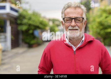 Felice bello senior barbuto uomo sorridente indossando occhiali e giacca rossa all'aperto Foto Stock