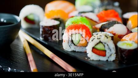 Diversi tipi di sushi disposti su un piatto nero con bacchette e salsa di soia Foto Stock