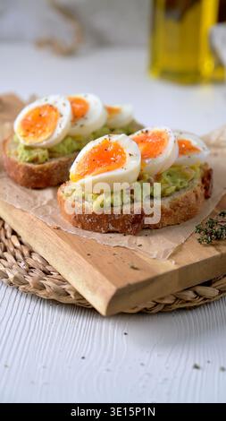 Due fette di pane a lievitazione naturale croccante ricoperte di purè di avocado e uova sode perfettamente morbide. Scatto ad angolo alto con luce naturale soffusa Foto Stock
