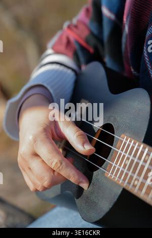 Primo piano di mani femminili che suonano la chitarra ukulele e cantano rilassandosi all'aperto nella soleggiata giornata autunnale. Donna hippie in allenamento in stile boho che suona strumento musicale. L'autunno lascia il clima autunnale Foto Stock
