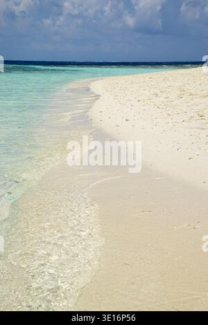 Le dolci onde dell'oceano raggiungono una spiaggia tropicale sabbiosa con acque turchesi poco profonde sotto un cielo nuvoloso. Foto Stock