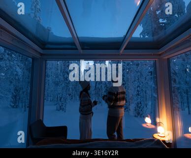 Due persone si godono un momento di pace all'interno di un'accogliente cabina di vetro, osservando il tranquillo paesaggio innevato di Akaslompolo, Lapponia. Il soffice bagliore delle lanterne crea un'atmosfera calda. Foto Stock
