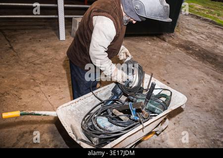 Un lavoratore che indossa indumenti protettivi organizza gli attrezzi elettrici e i cavi in una carriola in un cantiere, preparandosi per la manutenzione o la riparazione Foto Stock