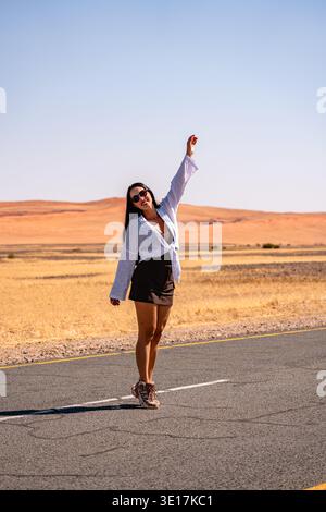 Una donna con gli occhiali da sole si erge gioiosamente su Sesriem Road a Sossusvlei, in Namibia. L'arido paesaggio desertico sullo sfondo cattura la serena bellezza Foto Stock