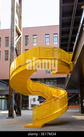 Una scala a chiocciola gialla in un ambiente industriale si erge sullo sfondo di un'architettura moderna. I colori vibranti contrastano con le travi metalliche Foto Stock