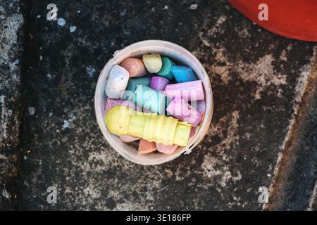 Vista dall'alto dei colorati pezzi di gesso del marciapiede in un contenitore di plastica su cemento grezzo all'aperto, vivaci tonalità pastello che contrastano con il surf intempestivo Foto Stock