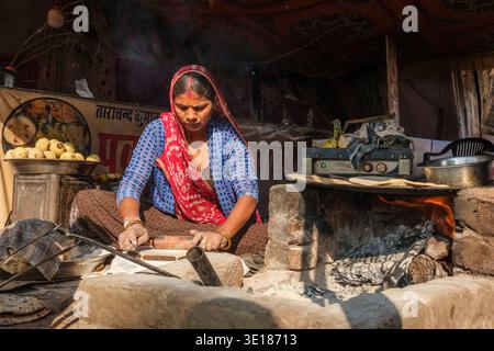 Una donna prepara la focaccia tradizionale sopra un caminetto aperto in una bancarella di cibo di Pushkar Mela, con pile di spuntini cotti visibili in lontananza. Foto Stock