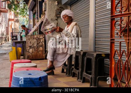 Un uomo che indossa un turbante bianco si siede su uno sgabello di plastica in un vicolo tranquillo di Pushkar mentre legge un giornale di fronte alle persiane chiuse. Foto Stock