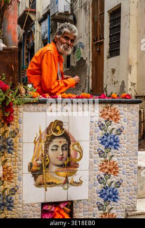 Un uomo dai capelli grigi con una camicia arancione siede a Pushkar dietro un muro piastrellato che raffigura la divinità Shiva, con un vicolo stretto dietro. Foto Stock