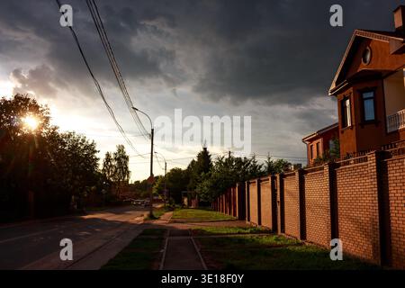 Contrasto spettacolare in una strada suburbana. Il sole che sbircia tra gli alberi illumina un muro di mattoni e una casa, mentre le nuvole di tempesta scure si stagliano sopra la testa Foto Stock