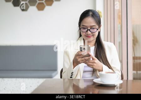 Giovane donna d'affari asiatica seduta nel ristorante caffetteria con indosso abiti e occhiali da vista mentre usa lo smartphone Foto Stock