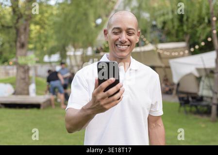 Elegante uomo calvo con una polo bianca che si rilassa in campeggio accanto a un camper. Viaggi all'aperto e concetto di stile di vita per il campeggio con un ambiente erboso Foto Stock