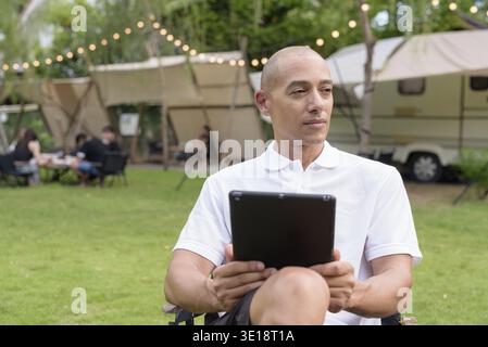 Elegante uomo calvo con una polo bianca che si rilassa in campeggio accanto a un camper utilizzando un tablet. Concetto di stile di vita per viaggi all'aperto e campeggio Foto Stock