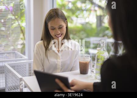 Due donne asiatiche hanno una piacevole conversazione con un caffè in un moderno ambiente da caffe' con vegetazione esterna Foto Stock