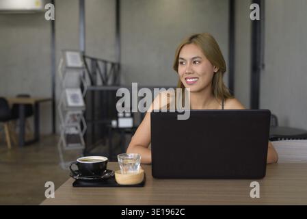 Ritratto di una bella giovane donna asiatica seduta in un bar che si rilassa e indossa abiti professionali mentre usa il computer portatile. Gli indumenti indicano il bus Foto Stock