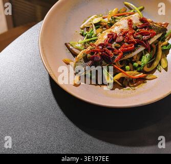 Filetto di spigola grigliato servito su un letto di verdure saltate, condite con erbe e spezie, creando un piatto visivamente attraente e saporito Foto Stock