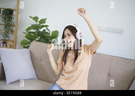 Felice giovane donna asiatica che indossa le cuffie divertirsi e ballare ascoltando la musica rilassarsi nel soggiorno. bella donna gioiosa che balla in casa Foto Stock