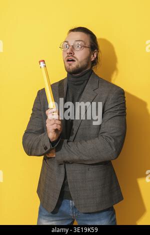 Uomo barbuto ispirato che indossa occhiali con una matita grande su sfondo giallo Foto Stock