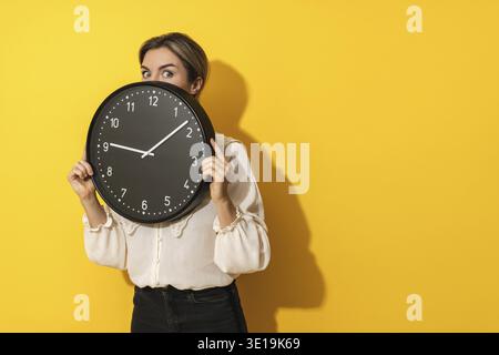 Giovane donna d'affari che sbircia da dietro un grande orologio su sfondo giallo Foto Stock