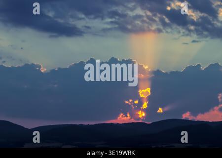 Potenti raggi solari scoppiano verso l'alto da dietro nuvole scure, proiettando un bagliore viola e arancione sull'orizzonte della montagna. Foto Stock
