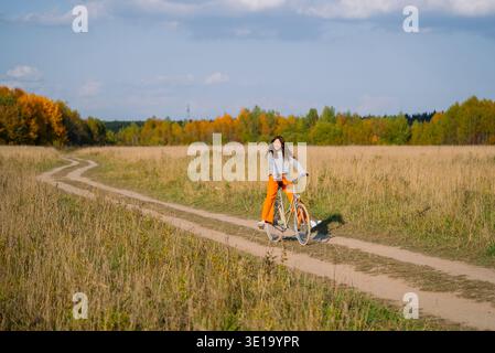 Giovane donna sorridente, che si gode un giro in bicicletta attraverso un campo d'oro autunnale su un tortuoso sentiero sterrato, abbracciando la libertà e uno stile di vita sano all'aria aperta nella tranquilla campagna Foto Stock