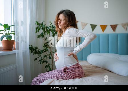 Donna incinta seduta a letto a casa, che stringe la schiena e soffre di disagio, soffre di mal di schiena e affaticamento durante la maternità Foto Stock
