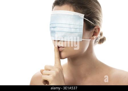 La giovane donna indossa una maschera di prevenzione sugli occhi facendo gesti di shush. Concetti di teoria della cospirazione o disinformazione sulla pandemia di COVID-19 Foto Stock