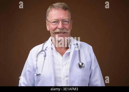 Foto in studio di un bellissimo medico anziano con baffi che indossano occhiali da vista su sfondo marrone Foto Stock