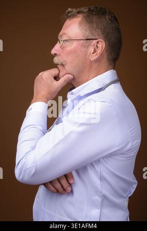 Foto in studio di un bellissimo medico anziano con baffi che indossano occhiali da vista su sfondo marrone Foto Stock