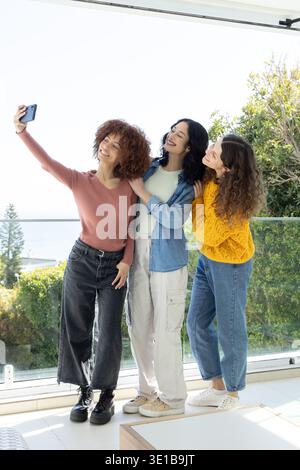 Diverse amiche in posa per un selfie su un balcone di vetro che si affaccia sull'acqua, tenendo in mano lo smartphone Foto Stock