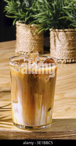 Un bicchiere di caffè ghiacciato è seduto su un tavolo da caffè al sole, con due piante sullo sfondo fuori fuoco Foto Stock