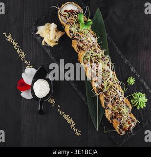 Il fresco rotolo di sushi tempura maki si trova su un piatto di ardesia nero decorato con fiori e semi di sesamo Foto Stock
