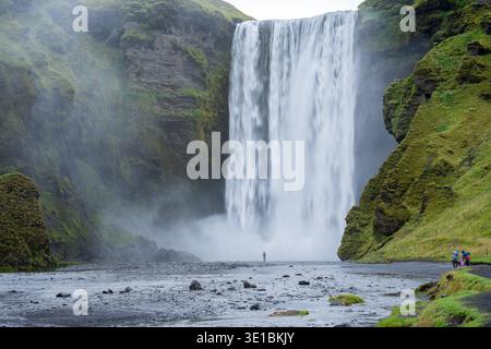 Enorme cascata circondata da scogliera rocciosa rocciosa e persone che camminano verso di essa, l'Islanda Foto Stock