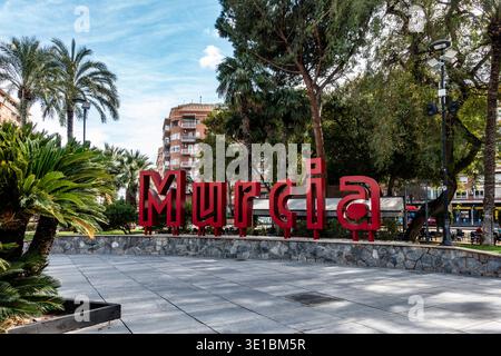 Grande cartello rosso di Murcia in una piazza lastricata a Murcia, Spagna. Circondato da palme, vegetazione ed edifici residenziali sotto un cielo blu. Foto Stock