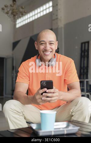 Bell'uomo calvo di mezza età con una polo arancione al tavolino da caffè con una tazza di caffè, cuffie e smartphone. Tecnologia e stile di vita c Foto Stock
