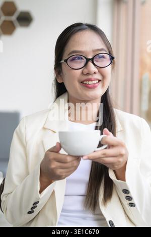 Giovane donna d'affari asiatica seduta in un ristorante caffetteria che indossa abiti e occhiali da vista e beve caffè Foto Stock