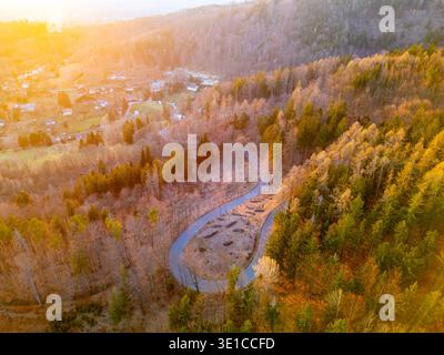 Una strada tortuosa attraversa una foresta di alberi in tonalità autunnali durante il tramonto. I colori vivaci illuminano il paesaggio, creando un'atmosfera tranquilla e serena. Foto Stock