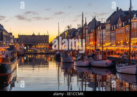 Mentre la sera scende su Nyhavn a Copenaghen, i colorati edifici illuminano il lungomare. Le barche si spostano dolcemente nel canale, mentre la gente si gode l'atmosfera vivace di accoglienti caffetterie. Foto Stock