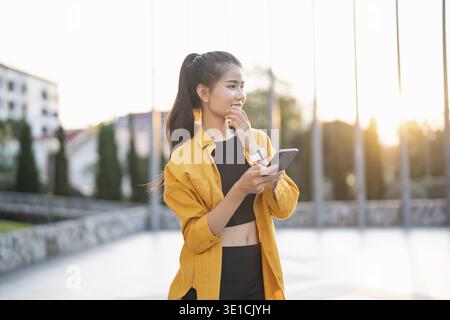 Sorridente giovane donna asiatica che usa lo smartphone in città controllando app mobili all'aperto Foto Stock