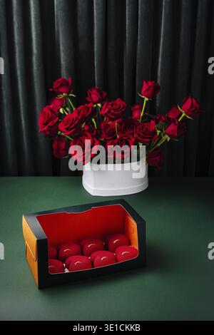 Un bellissimo display presenta rose rosse vivaci in un vaso bianco posto vicino a una scatola colorata che contiene piccoli oggetti decorativi. Lo sfondo scuro migliora la qualità Foto Stock