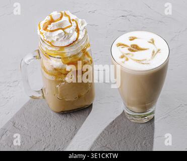 Due bevande a base di caffè sono presentate su una superficie ruvida. Una bevanda è in un barattolo con panna montata e gocciolamento di caramello, mentre l'altra è una schiuma Foto Stock