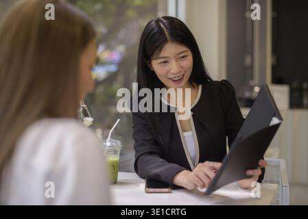 Due donne asiatiche hanno una piacevole conversazione con un caffè in un moderno ambiente da caffe' con vegetazione esterna Foto Stock