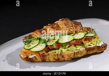 Elegante panino con croissant con salmone affumicato, avocado, cetrioli ed erbe fresche su un piatto bianco Foto Stock