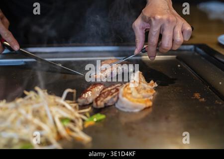 Le mani dello chef con la spatola sopra il teppanyaki. cucinare verdure, carne e pesce su un tavolo caldo per grigliate hibachi. Cucina tradizionale giapponese. Spettacolo Teppan Foto Stock