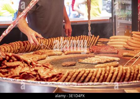 Ampia griglia ripiena di vari tipi di salsicce, polpettine e carne cucinata. Una persona tende a grigliare, utilizzando pinze per preparare la salsiccia. La S Foto Stock