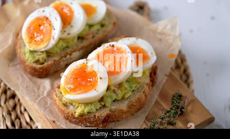 Due fette di lievito madre croccante ricoperte di crema di avocado e uova sode soffici. La scena è caratterizzata da un'illuminazione naturale brillante e rustica Foto Stock