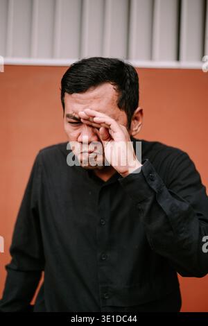 Un uomo con una camicia nera si strofina gli occhi, esprimendo disagio, stanchezza o mal di testa, probabilmente a causa dello sforzo oculare degli schermi o della mancanza di sonno. Foto Stock